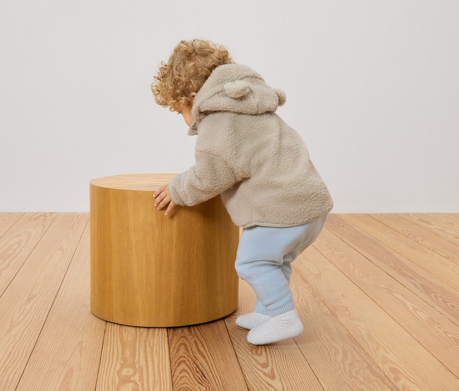 Un jeune enfant aux cheveux bouclés, vêtu d'un manteau teddy beige à capuche avec des oreilles, s'appuie sur un tabouret rond en bois.