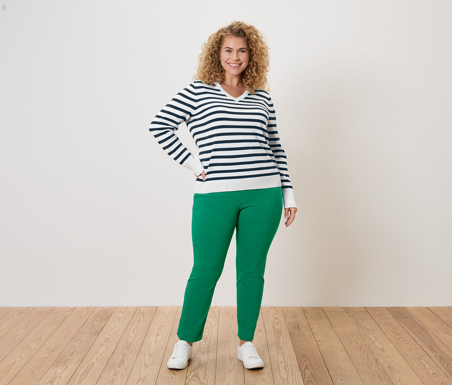 Une femme aux cheveux bouclés, portant un pull rayé bleu et blanc, un pantalon vert et des baskets blanches, pose sur un fond blanc.