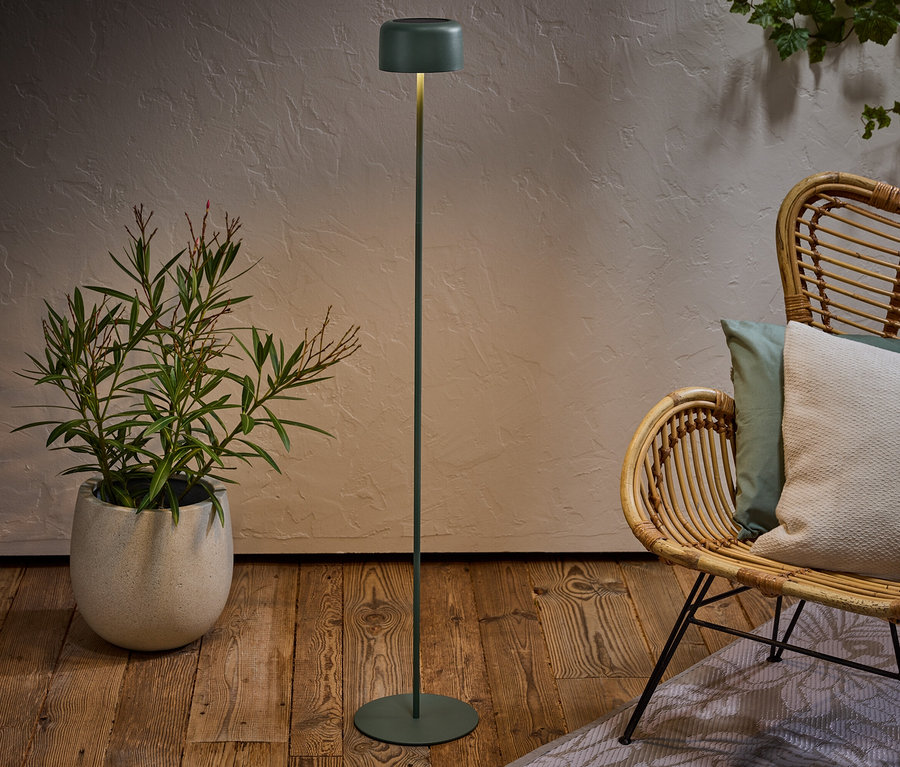 Un lampadaire vert se dresse à côté d'un fauteuil en rotin avec des coussins et d'une plante en pot sur un plancher en bois.