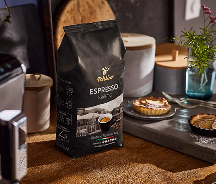 Un paquet de café en grains Tchibo Espresso Kräftig est posé sur une table en bois à côté d'une machine à café et d'assiettes avec des tartelettes.