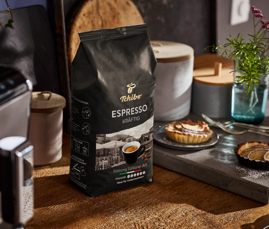 Un paquet de café en grains Tchibo Espresso Kräftig est posé sur une table en bois à côté d'une machine à café et d'assiettes avec des tartelettes.