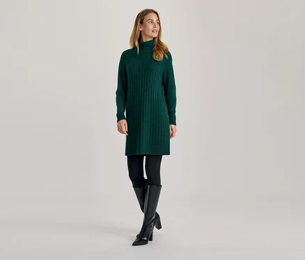 Femme portant une robe pull verte à col roulé, des leggings noirs et des bottes noires montantes, regardant sur le côté.