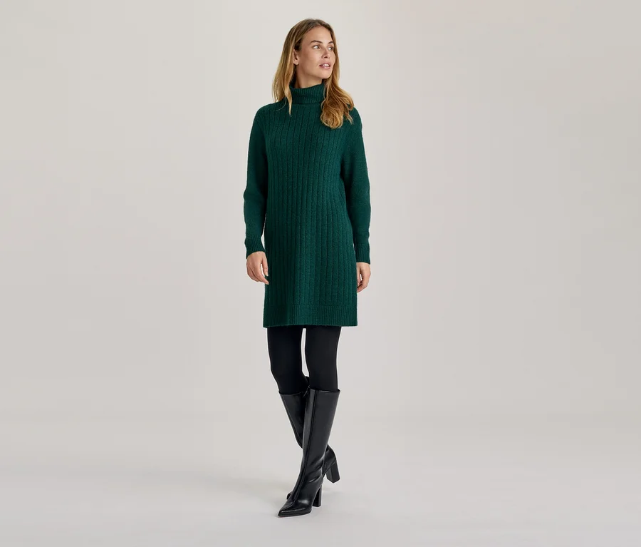 Femme portant une robe pull verte à col roulé, des leggings noirs et des bottes noires montantes, regardant sur le côté.