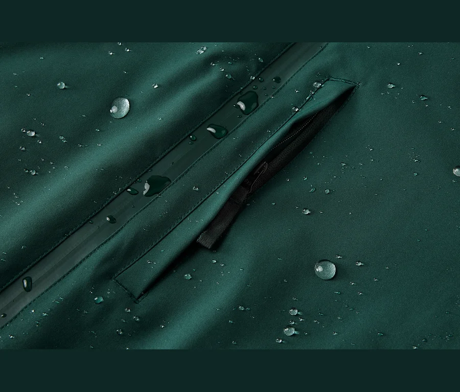 Gros plan d'un tissu imperméable vert foncé avec de nombreuses gouttelettes d'eau perlées à la surface, montrant une poche zippée noire et une couture scellée.