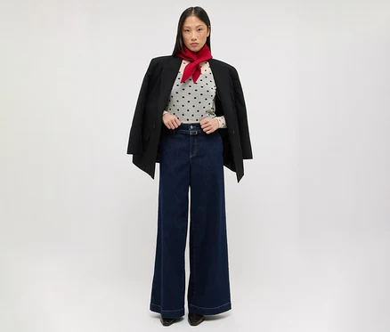 Une femme est debout, vêtue d'un blazer noir drapé sur ses épaules, d'un haut clair à pois, d'un jean large bleu foncé et d'un foulard rouge autour du cou.