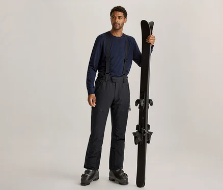 Un homme aux cheveux bouclés foncés et à la barbe claire est debout, portant un t-shirt à manches longues bleu foncé, un pantalon de ski noir à bretelles et des chaussures de ski noires. Il tient une paire de skis noirs à la verticale avec sa main droite.