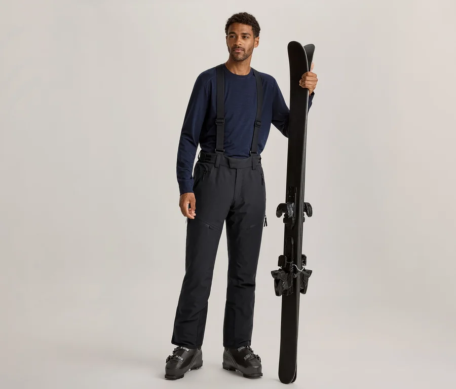 Un homme aux cheveux bouclés foncés et à la barbe claire est debout, portant un t-shirt à manches longues bleu foncé, un pantalon de ski noir à bretelles et des chaussures de ski noires. Il tient une paire de skis noirs à la verticale avec sa main droite.
