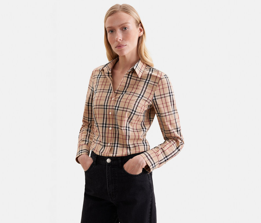 Femme blonde portant une chemise à manches longues à carreaux beige avec des lignes noires, rouges et blanches, et un jean noir, les mains dans les poches, devant un fond blanc.