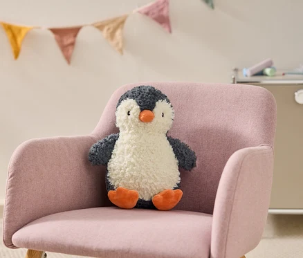 Une peluche pingouin duveteuse gris foncé et blanche avec un bec et des pattes orange est assise sur un fauteuil rose. Des guirlandes de fanions colorés sont accrochées en arrière-plan.