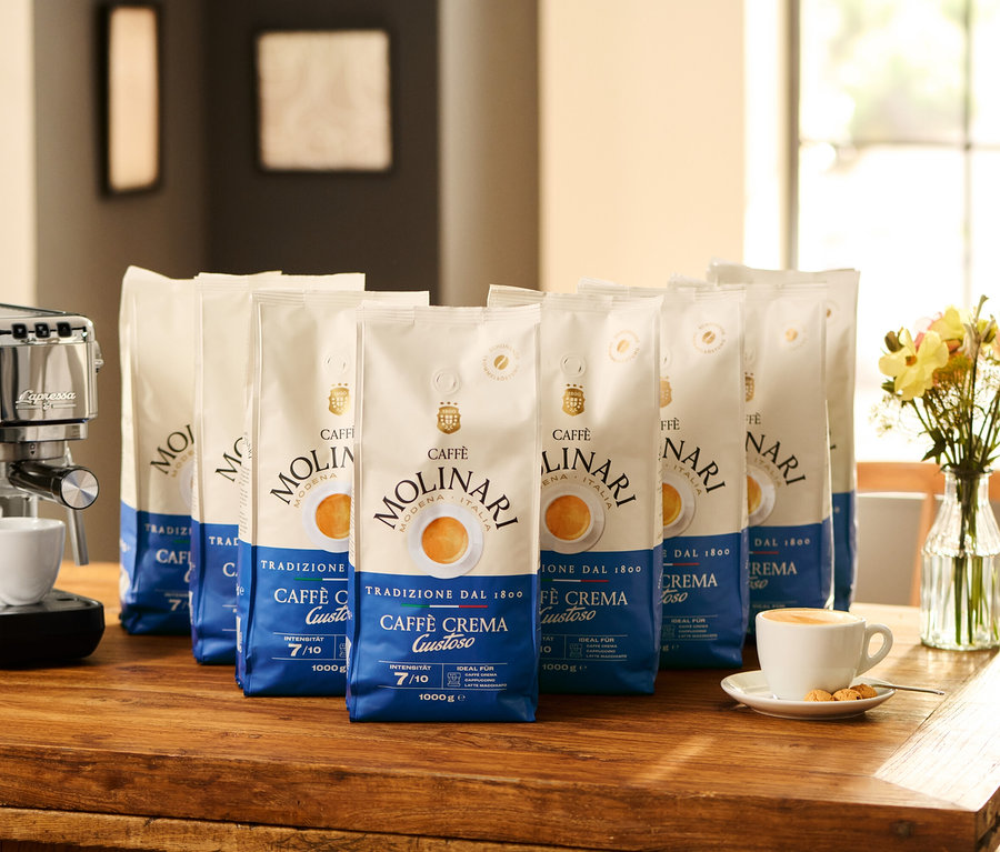 Machine à expresso avec tasse et huit paquets de Caffè Molinari Caffè Crema Gustoso en grains – 8x 1 kg sur une table en bois.