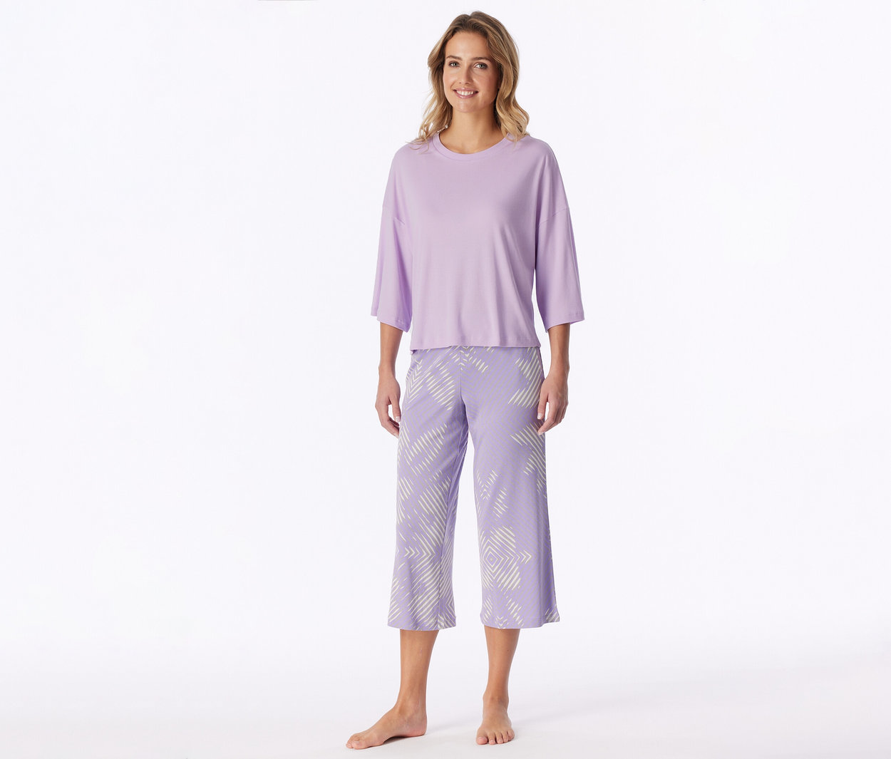 Femme souriante, pieds nus, portant un t-shirt lilas à manches trois-quarts et un pantacourt assorti à motif géométrique blanc.