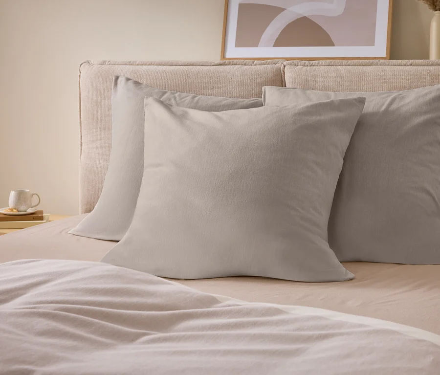 Un lit avec une tête de lit rembourrée beige, trois oreillers gris clair et une literie beige clair. Une tasse blanche sur des livres est posée sur une table de chevet en bois. Une œuvre d'art abstraite et des vases sont en arrière-plan.
