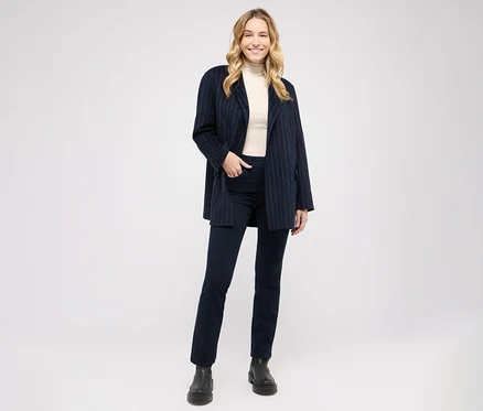 Une femme blonde souriante porte un blazer bleu marine à rayures, un col roulé crème, un jean foncé et des bottes noires.