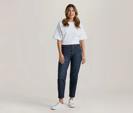 Femme debout et souriante, vêtue d'un t-shirt blanc à manches courtes, d'un jean bleu foncé et de baskets blanches.