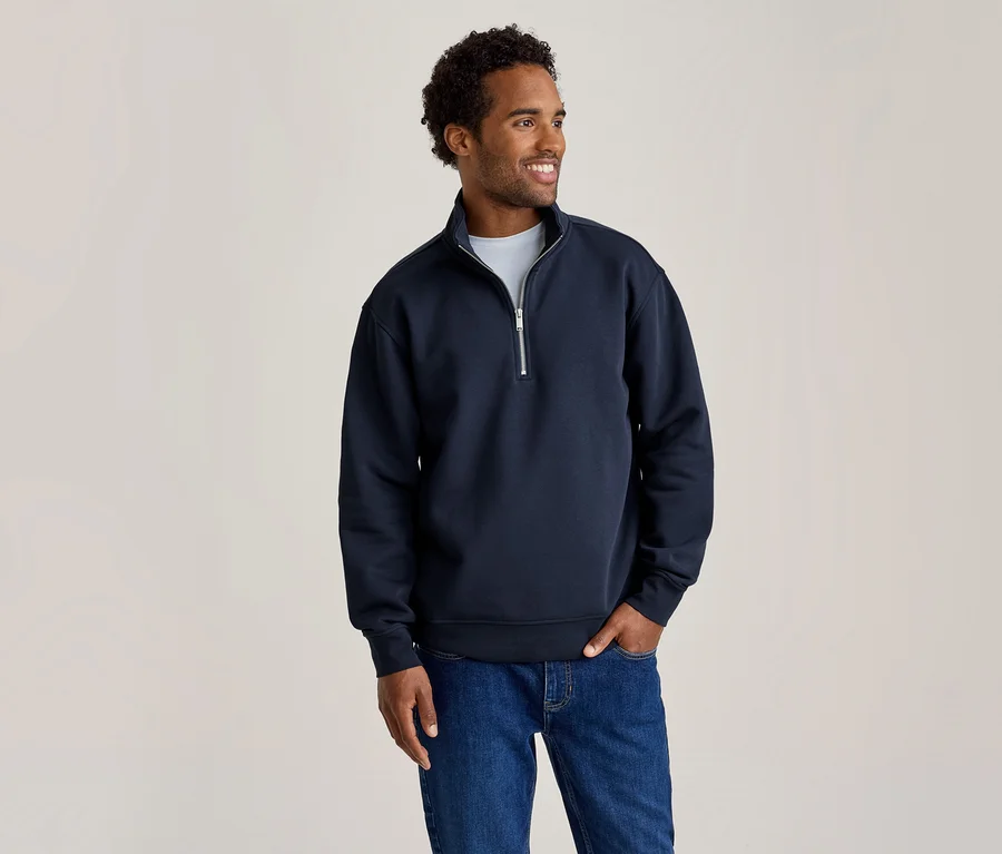 Homme portant un sweat-shirt bleu marine à col zippé et un jean bleu, souriant et regardant vers la droite, une main dans sa poche.