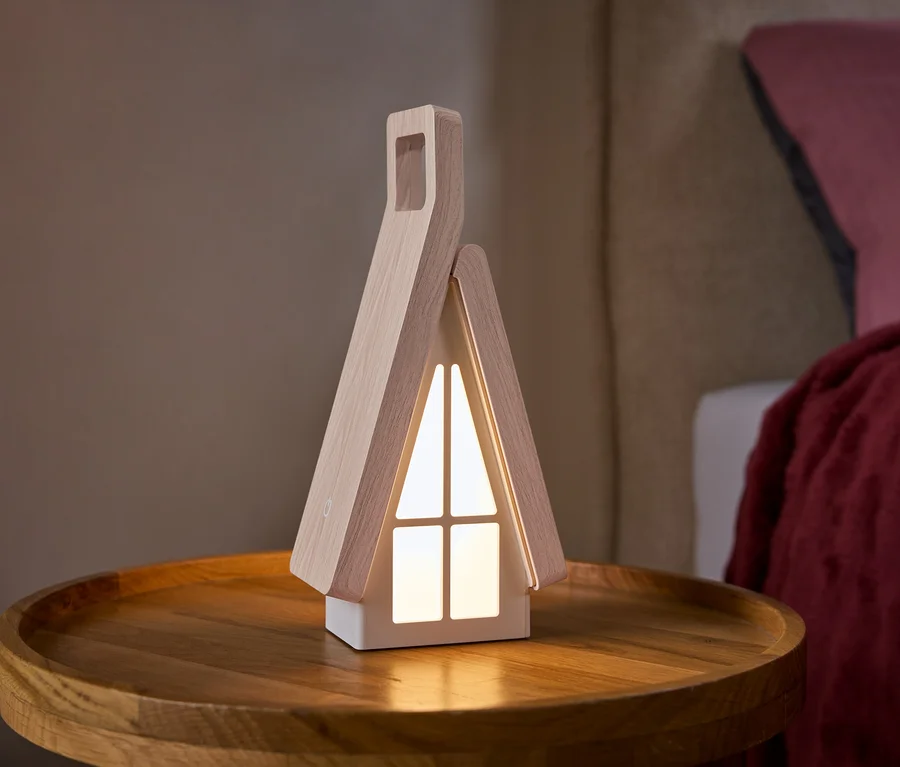 Une lampe en forme de maison avec des côtés en bois clair et des fenêtres blanches lumineuses est posée sur une table d'appoint ronde en bois. Un lit avec un oreiller rose et une couverture rouge est en arrière-plan.