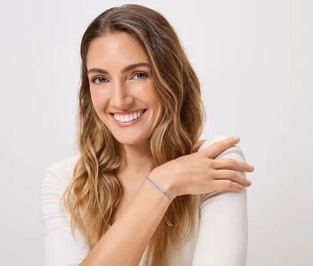 Femme souriante aux cheveux blonds ondulés portant un bracelet bleu au poignet gauche sur fond blanc.