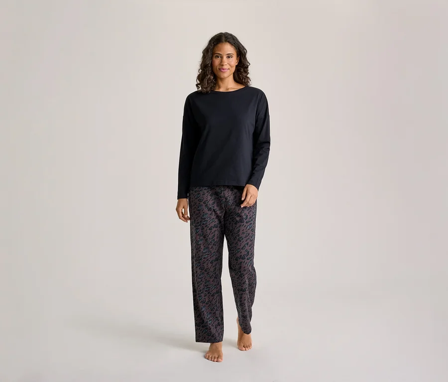 Une femme aux cheveux foncés et bouclés se tient de face, vêtue d'un haut noir à manches longues et d'un pantalon de pyjama foncé à motifs. Elle est pieds nus.