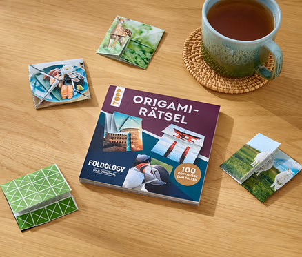 Sur une table en bois sont posés le livre »Origami-Rätsel«, une tasse de thé et des figurines origami pliées avec différents motifs.