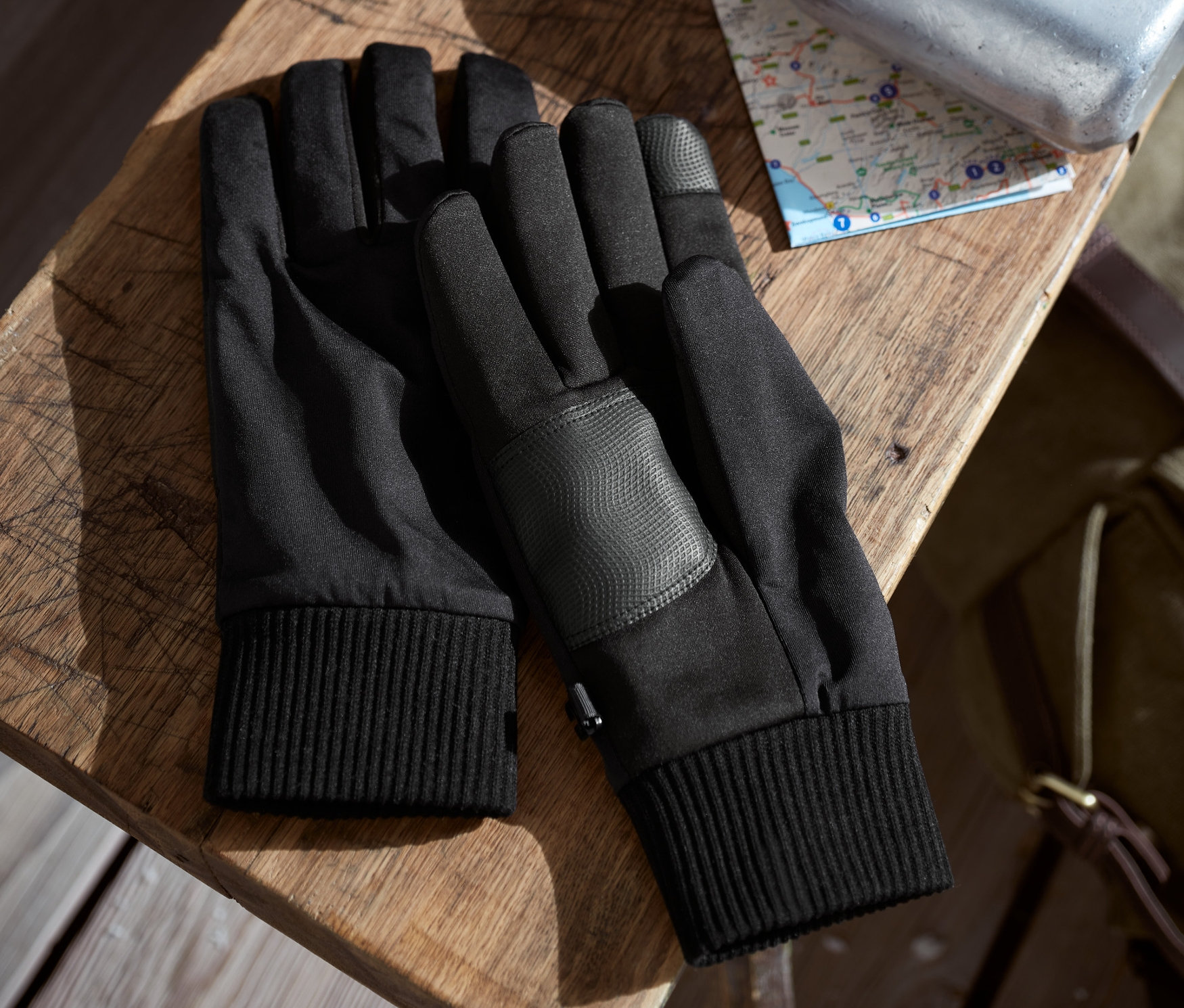 Deux paires de gants softshell noirs sont posées côte à côte sur une table en bois. Derrière, il y a une carte et une bouteille argentée.