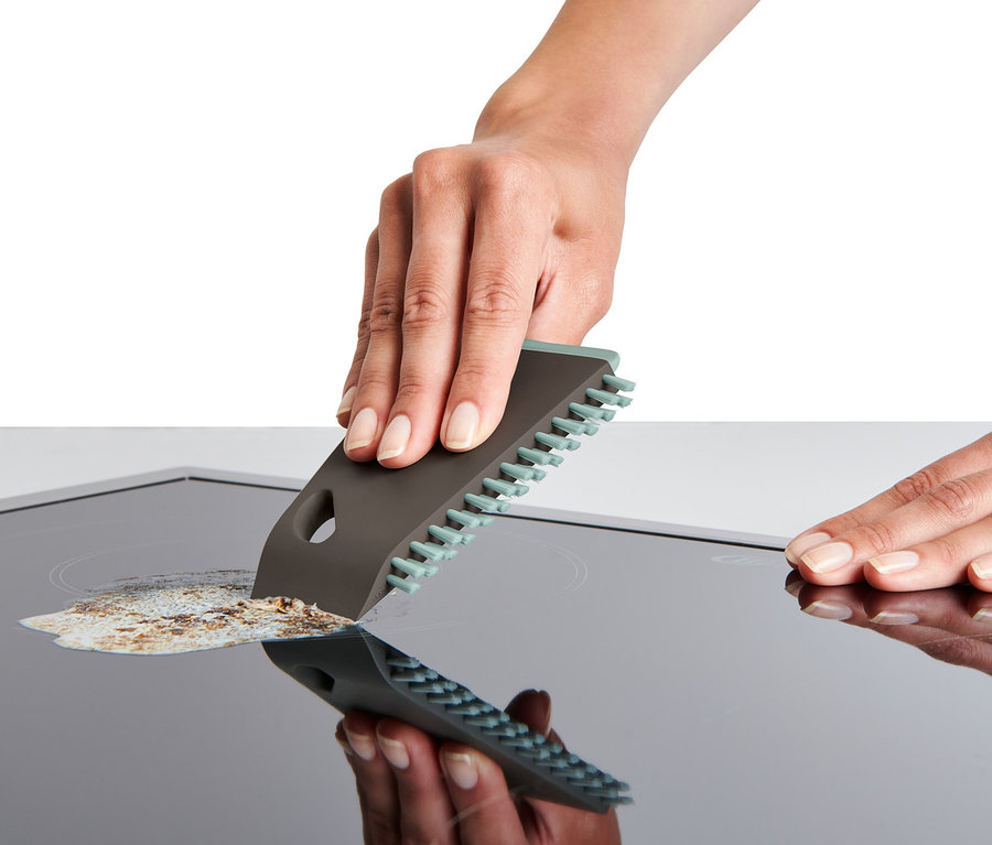 Une main nettoie une plaque vitrocéramique avec une brosse de cuisine universelle.