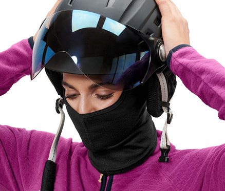 Une femme met un casque de ski noir avec visière et porte une cagoule performance noire.
