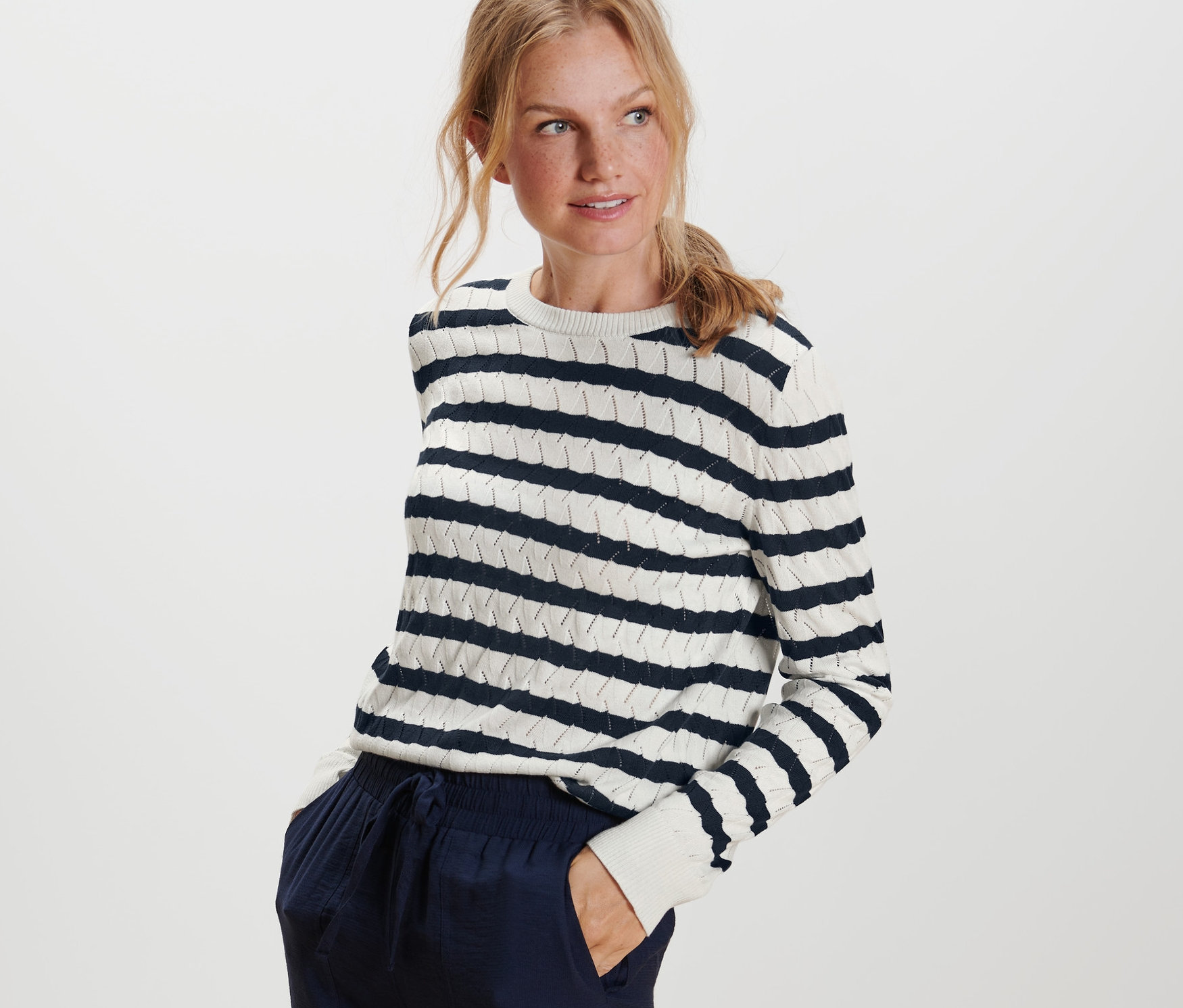 Une femme blonde pose, portant un pull en maille à rayures.