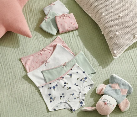 Sept culottes pour tout-petits sont posées sur un fond vert à côté d'un jouet et de coussins.