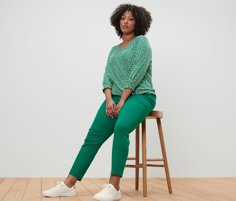 Une femme est assise sur un tabouret et porte une blouse imprimée verte et un pantalon stretch vert.