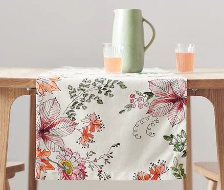 Sur une table en bois, un chemin de table à motifs floraux. Sur la table, un pichet et deux verres contenant un liquide orange.