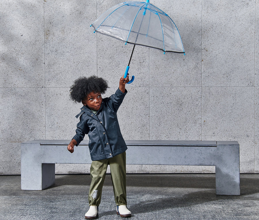 Garçon tenant un parapluie transparent bordé de bleu au-dessus de sa tête et portant une veste de pluie.