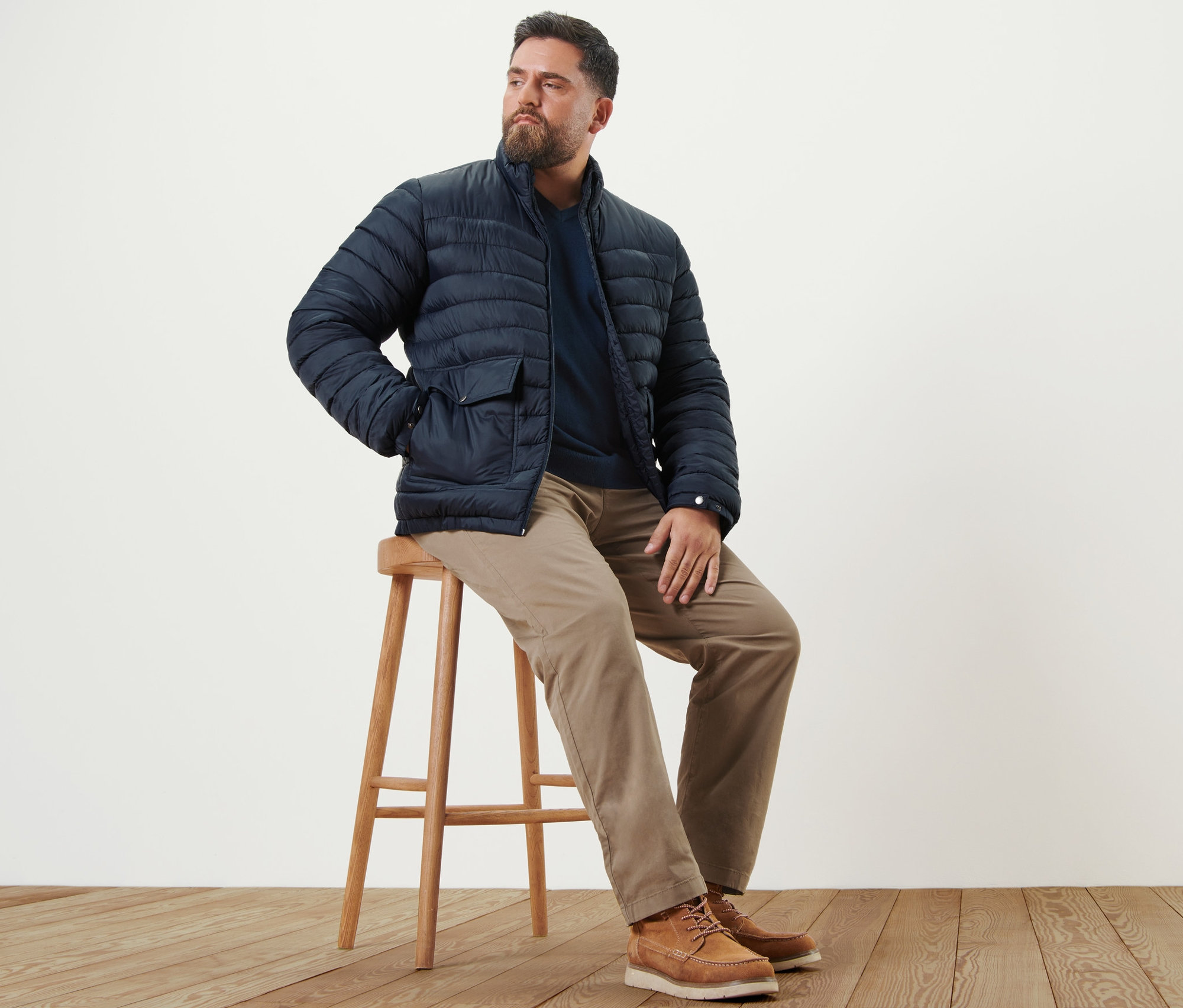 Un homme est assis sur un tabouret et porte un pantalon chino sable, un pull à encolure en V bleu marine et une veste matelassée bleu marine.