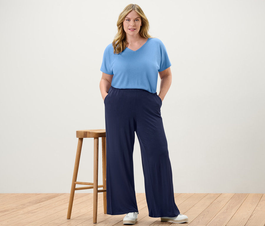 Une femme se tient debout, les mains dans les poches de son pantalon palazzo bleu foncé et d'un chemisier structuré bleu clair.