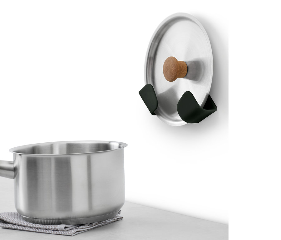 Une casserole en acier inoxydable avec une poignée en bois est posée sur un torchon gris. Au mur sont fixés 4 supports muraux pour couvercles.