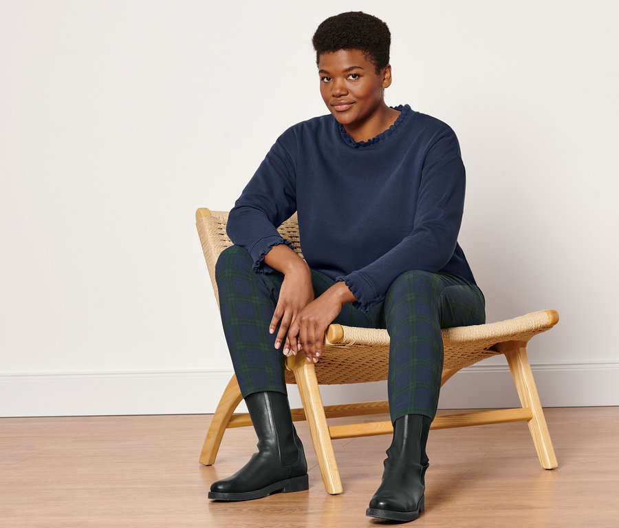 Une femme est assise sur une chaise en osier et porte un pantalon stretch à carreaux verts et bleus, des bottines en cuir et un sweatshirt avec des détails en tricot.