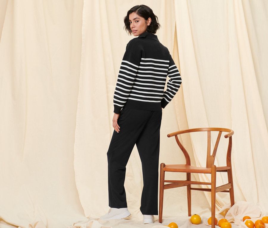 Une femme pose en pull avec col et un pantalon de jogging noir à côté d'une chaise en bois et des oranges.