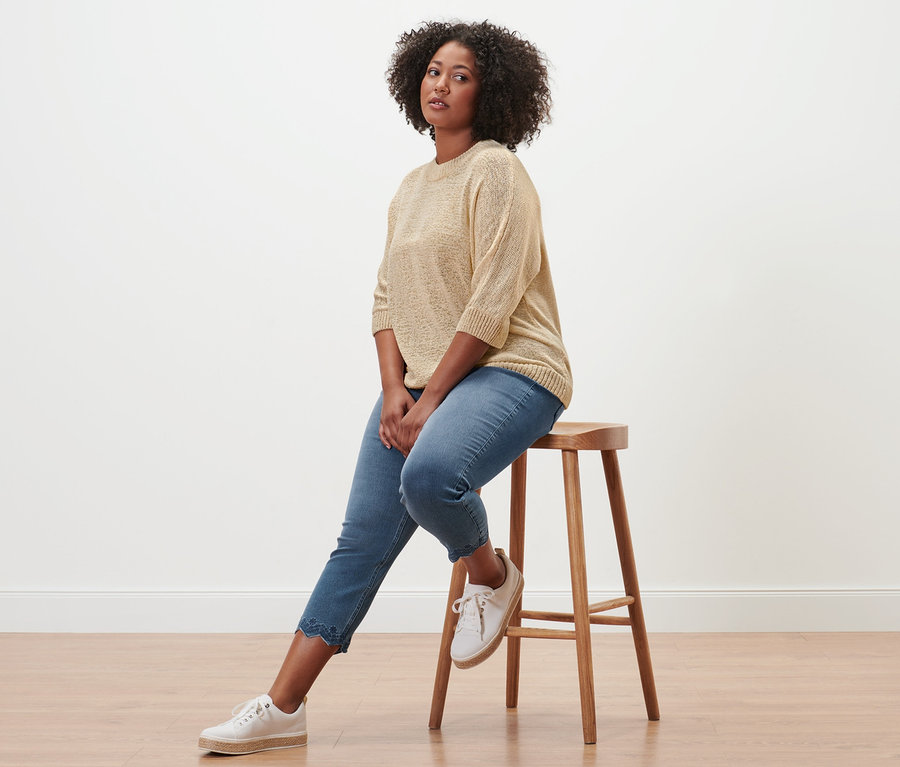 Une femme est assise sur un tabouret et porte un tregging 7/8 à broderie ajourée et un pull d’été en maille tricoté en fil à bandelettes.