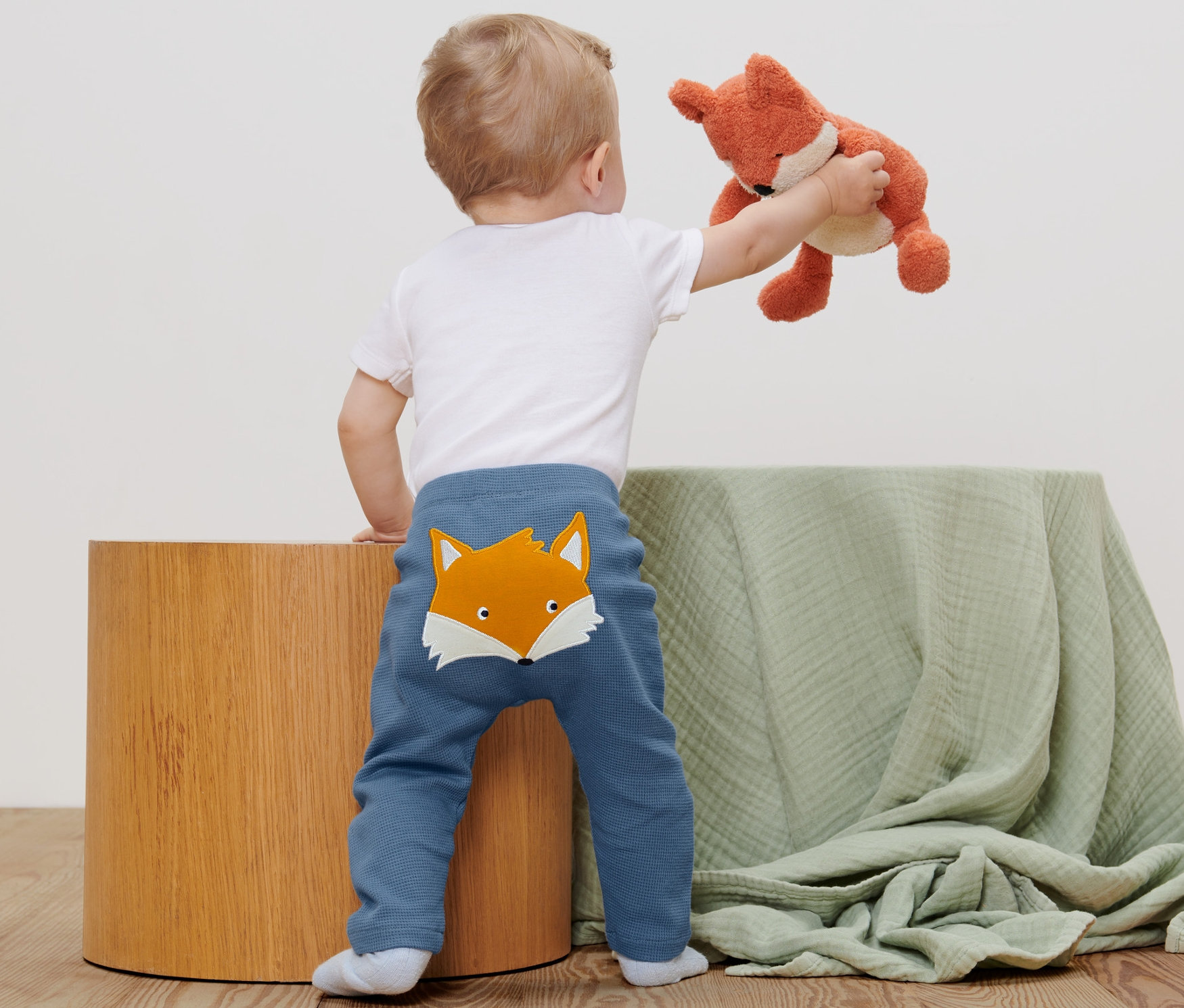 Un jeune enfant, vu de dos, tient un renard en peluche. Il porte 2 pantalons pour bébé.
