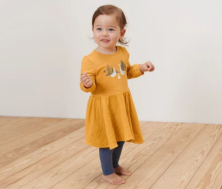 Une petite fille souriante se tient sur un plancher en bois et porte une robe tissée jaune pour bébé.