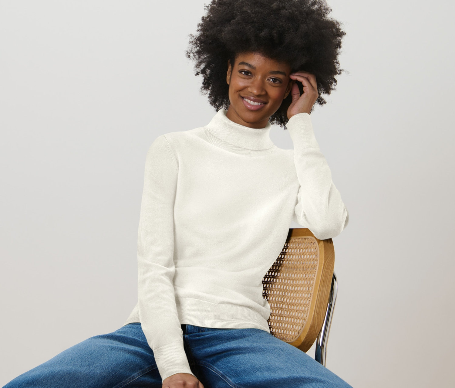 Une femme est assise sur une chaise et porte un pull à col roulé en maille fine blanc crème et un jean.