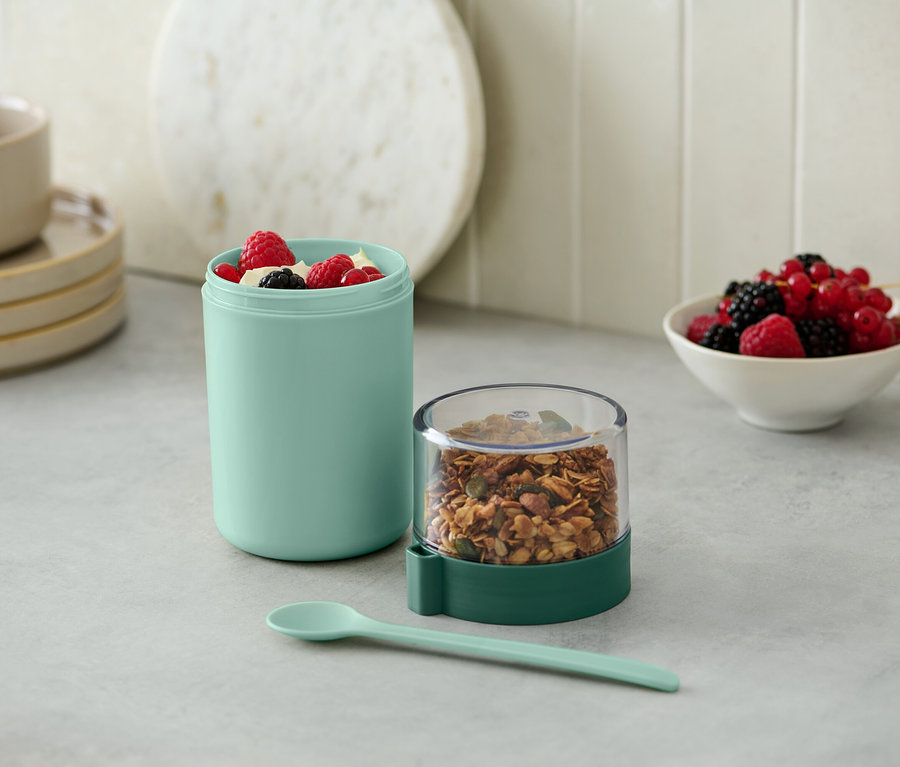 Sur une table, un récipient turquoise contenant des fruits et à côté un récipient transparent contenant du granola, le tout complété par une cuillère.
