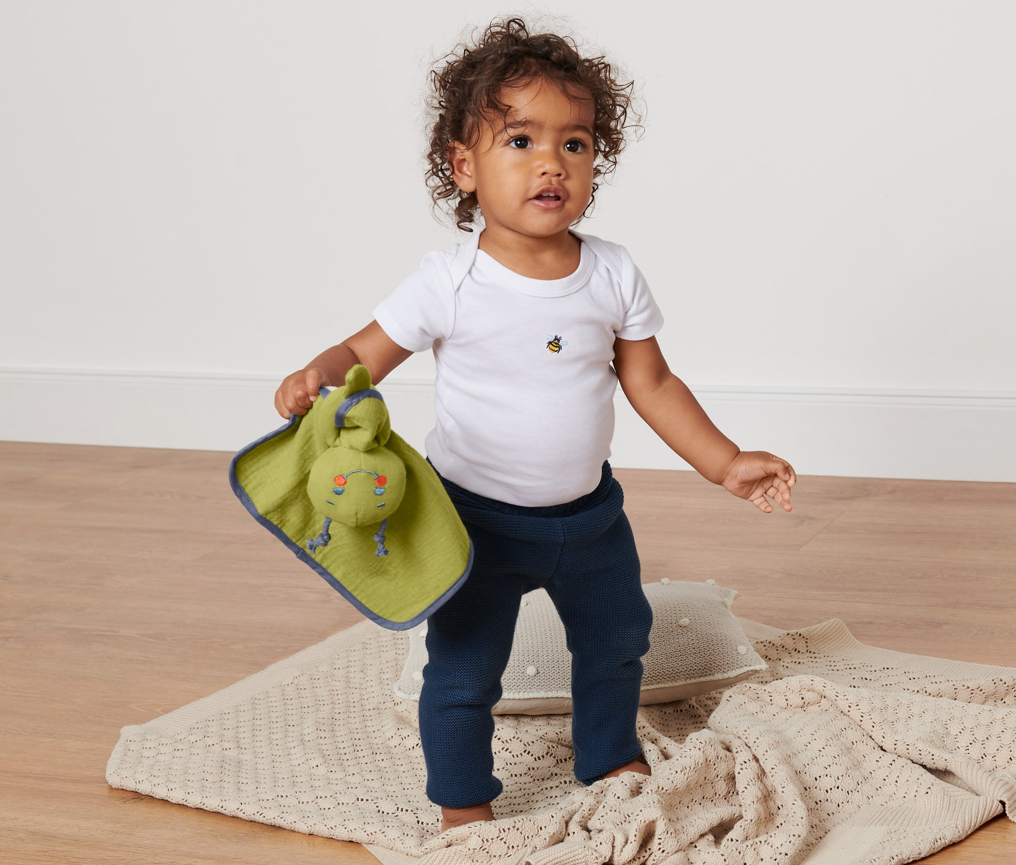 Un bébé se tient debout sur une couverture et tient un doudou en tissu pour bébé.