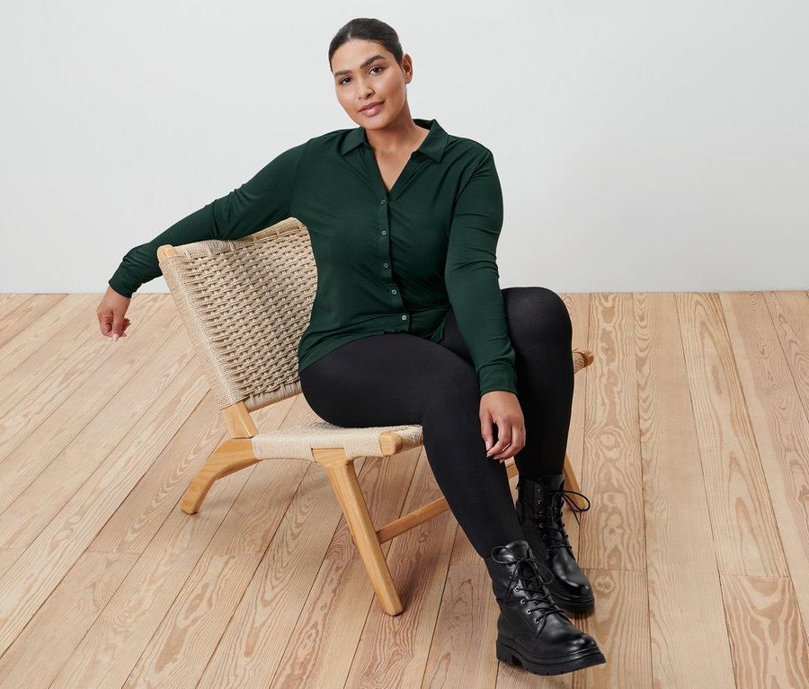 Une femme est assise sur une chaise en osier et porte une blouse vert foncé avec patte de boutonnage, un legging thermique avec relief et des bottines en cuir.