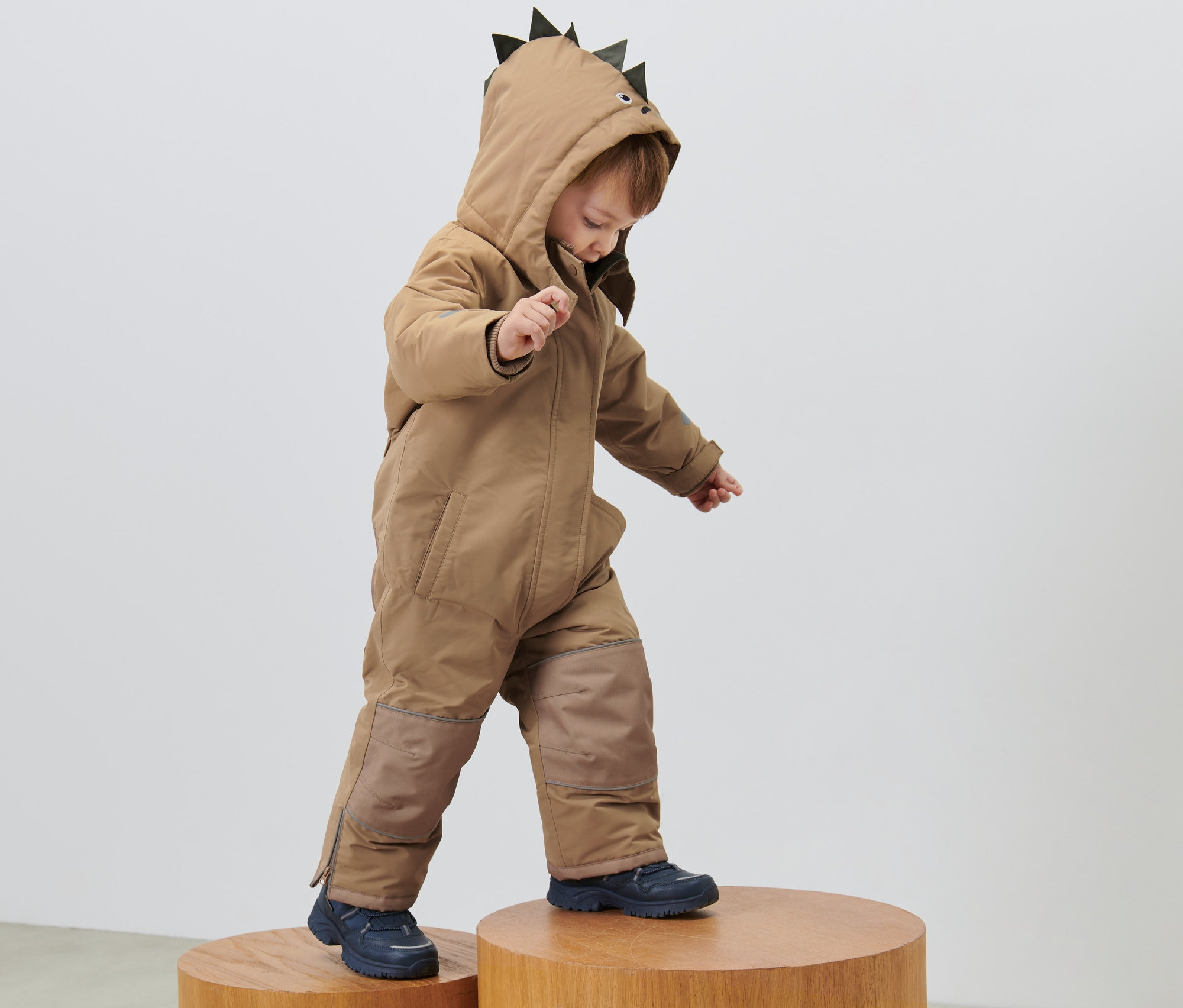 Un enfant dans une combinaison de neige pour enfant avec une capuche à pointes se tient sur des tabourets en bois.