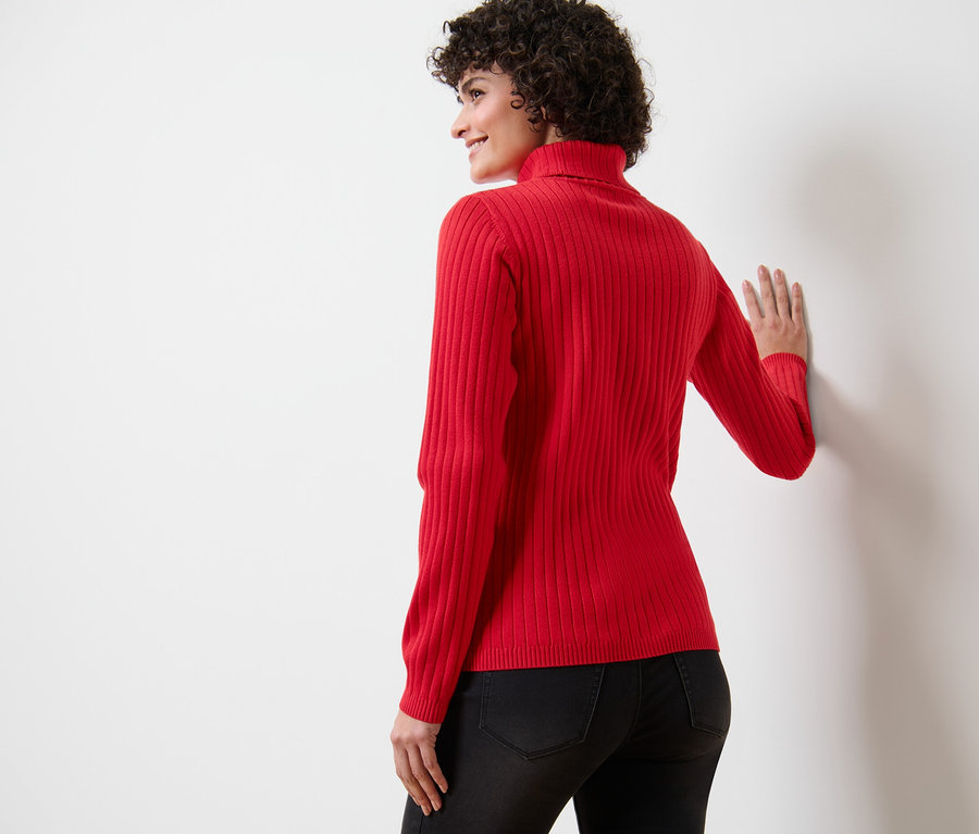 Femme appuyée contre un mur blanc portant un pull à col roulé côtelé rouge.