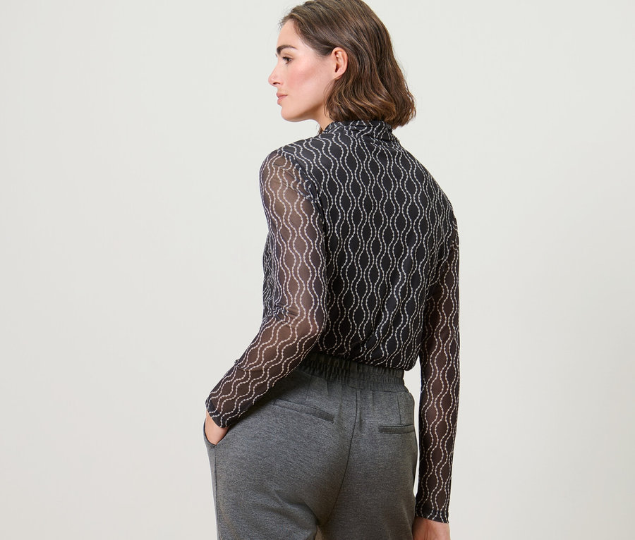 Femme debout, dos à la caméra, portant un haut manches longues à col roulé en mesh imprimé et un pantalon gris.