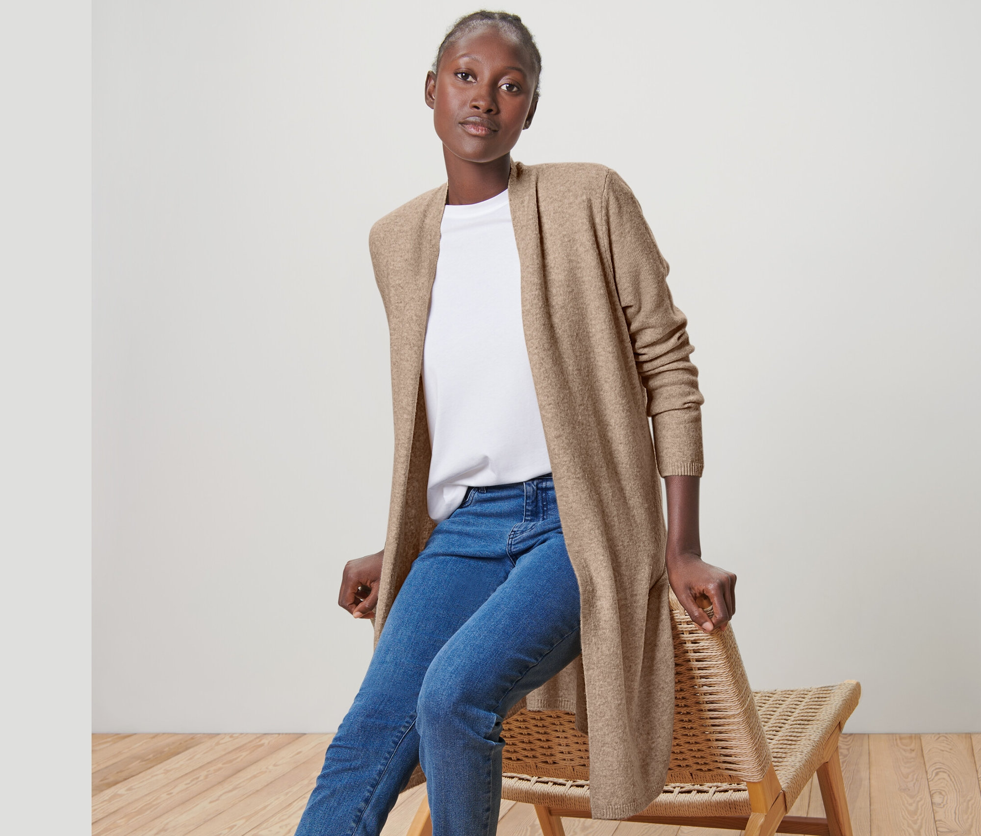 Une femme est assise sur une chaise et porte un cardigan long ouvert, un jean et des baskets.