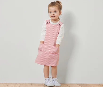 Fillette portant une robe-salopette tissée à rayures rouges sur un t-shirt blanc à manches longues.