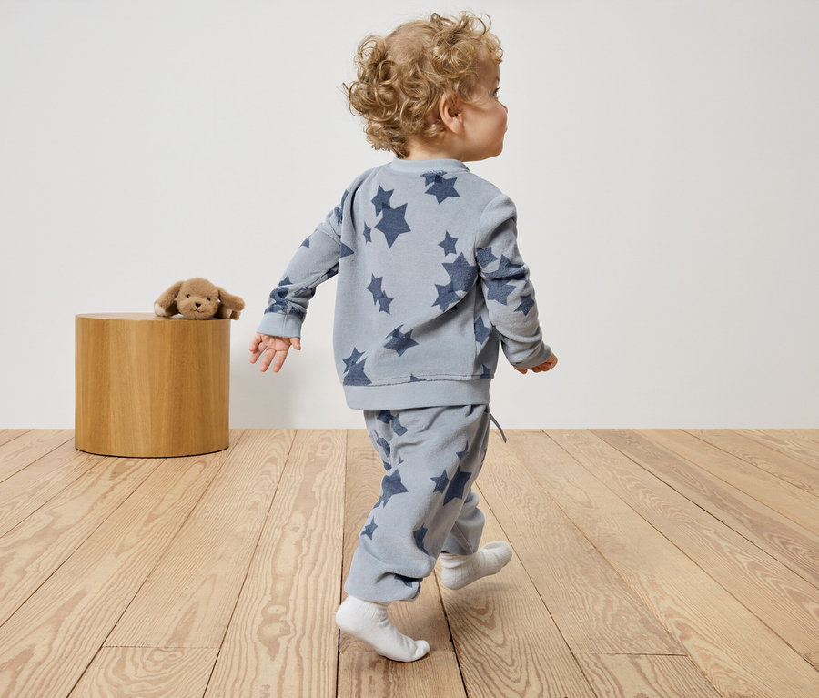 Bambin en vêtements bleus étoilés marchant sur un plancher en bois. Un chien en peluche est posé sur le tabouret.