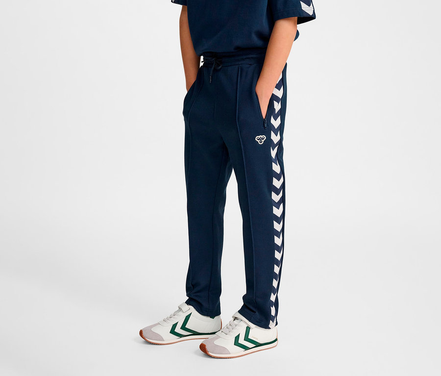 Un garçon debout porte un pantalon de survêtement bleu marine HUMMEL HMLJR Archive Regular Poly et des baskets.